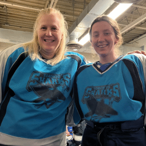 Author Sandra McTavish is dressed in a blue hockey jersey whose logo reads "Toronto Sharks". Her arm is draped over the shoulder of a fellow hockey player. Both of them smile widely at the viewer.