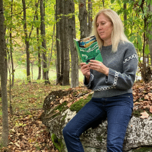 Author Sandra McTavish sits on a mossy rock in the middle of a deciduous forest. She holds a copy of The Cricket War open in front of her, and seems captivated by what she is reading.