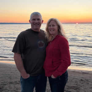 Author Sandra McTavish stands next to her partner Doug on a sandy beach. The sun sets on the horizon.